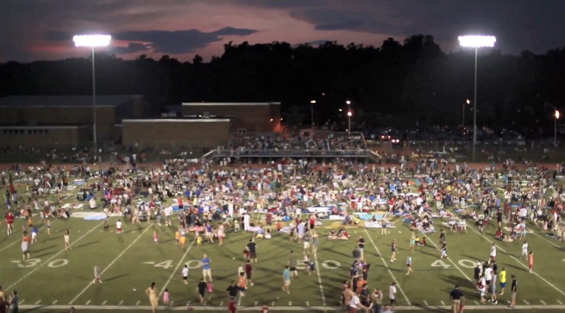 Families spread across the high school stadium field at dusk under the lights