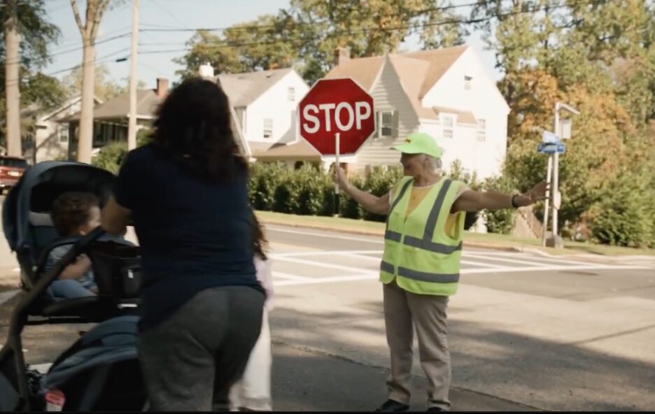 A crossing guard holding up a stop sign while an adult with a stroller and a child cross the street